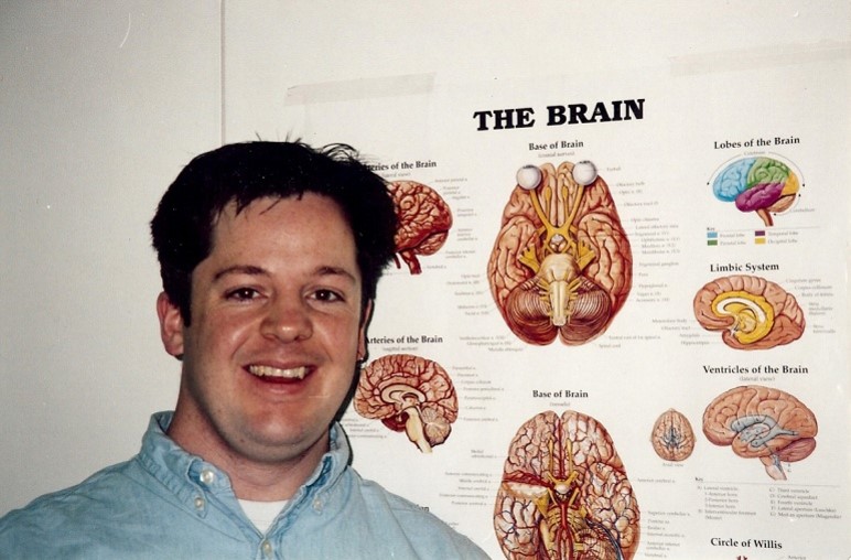 A smiling, young Andy Hill standing in front of an educational poster about the brain.