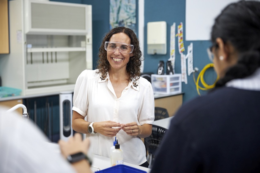 Photograph of a woman wearing safety glasses while in a lab. She is smiling while teaching students.